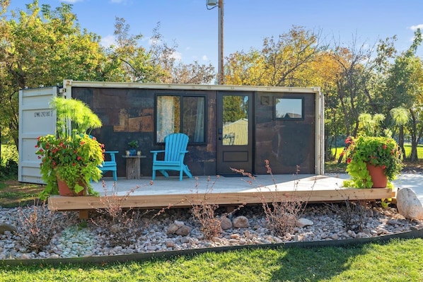 Une petite cabane moderne dotée d’une terrasse, de deux chaises et de plantes en pot.