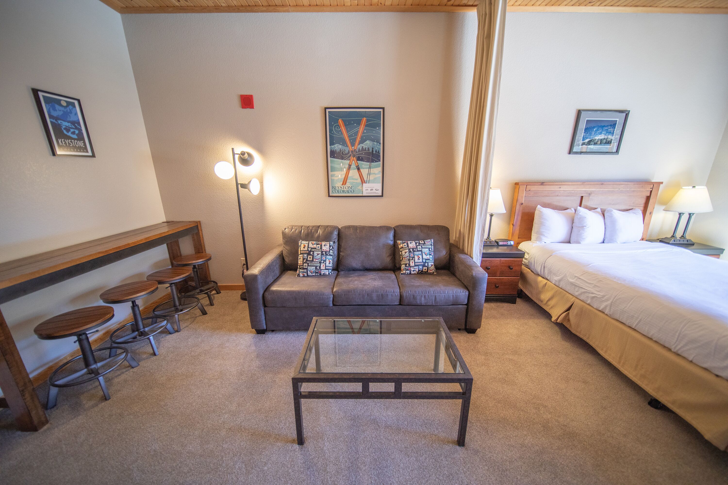 A hotel room with a sofa, a glass-top coffee table, a bed, a nightstand, a lamp, and a wall-mounted picture.