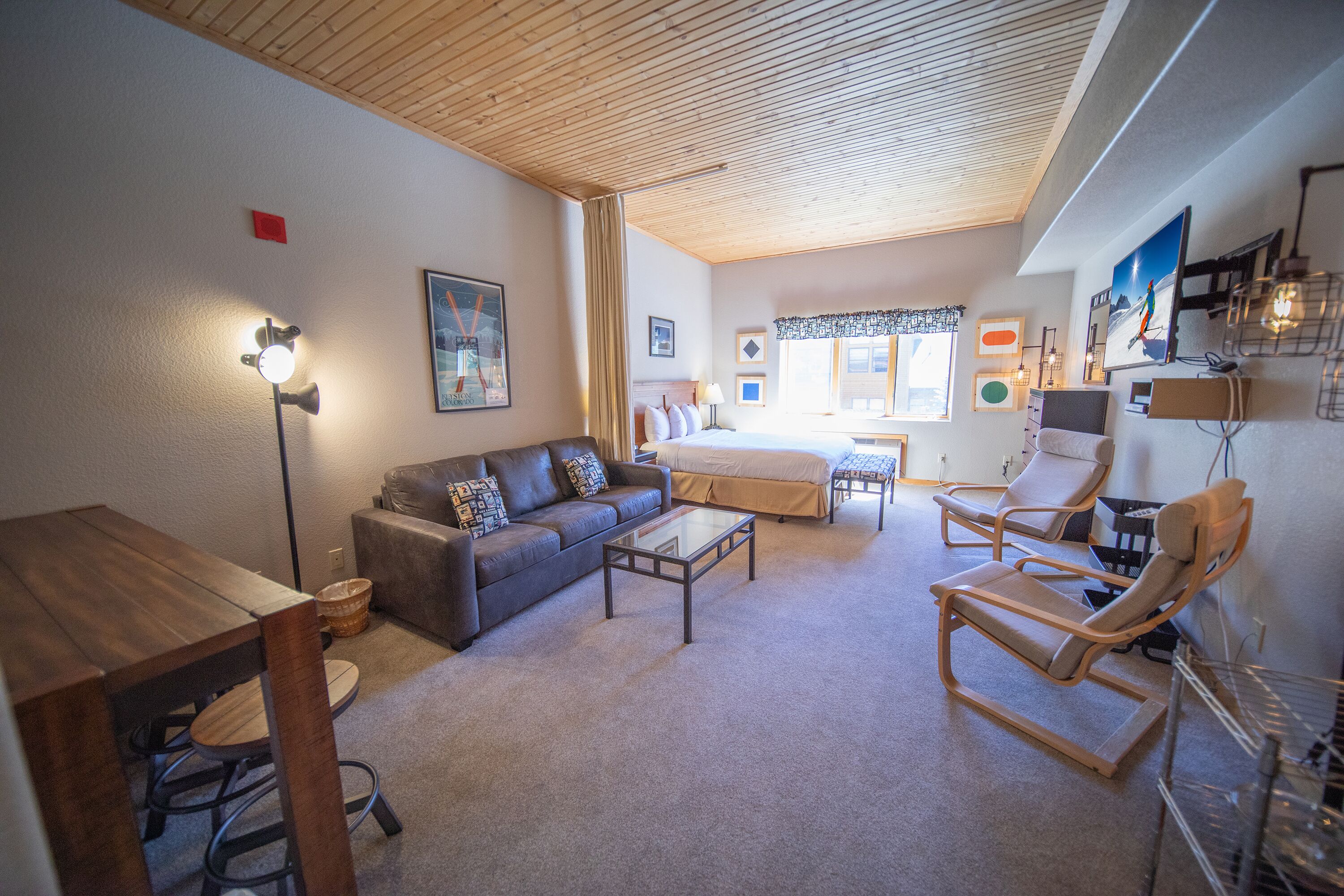 A cozy living room with a sofa, coffee table, and two lounge chairs. A bed is visible in the background.