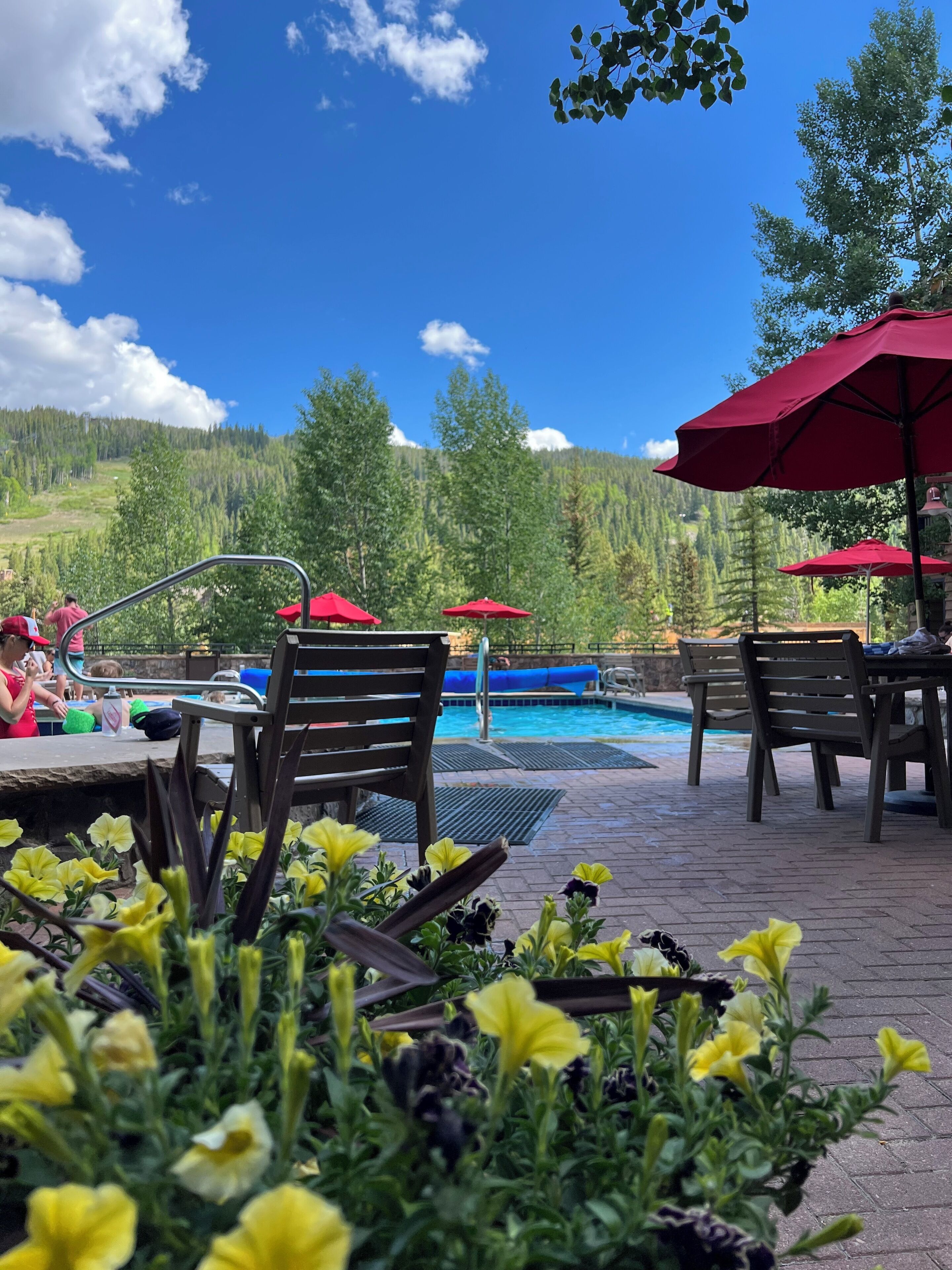 An outdoor pool area with red umbrellas, wooden chairs, and a view of a mountainous landscape.