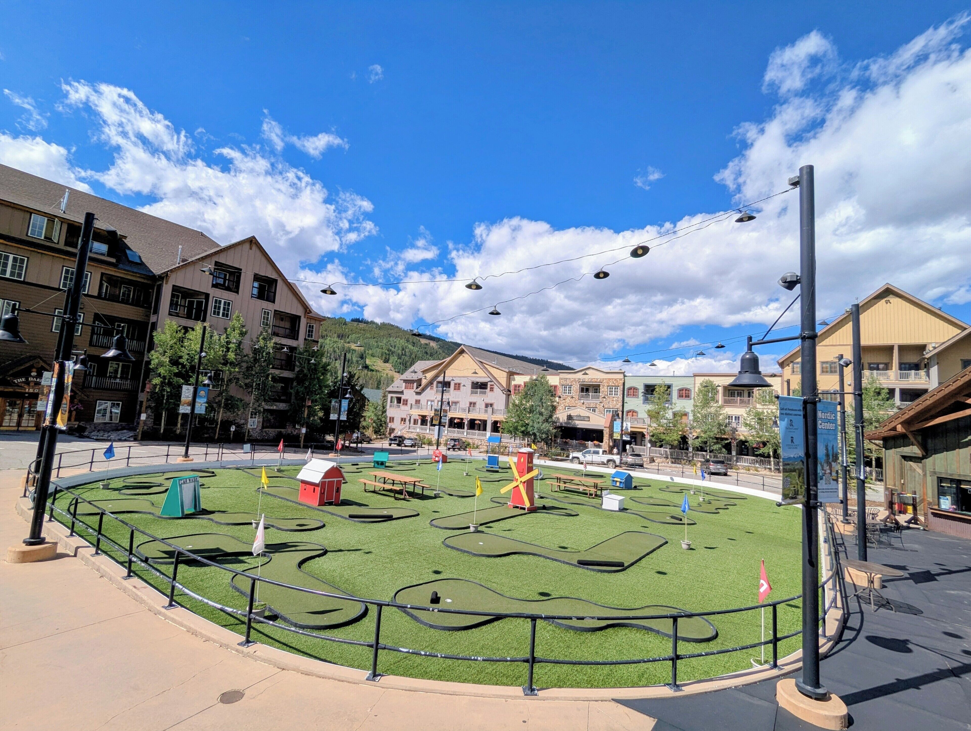 A landscaped area with a green lawn, benches, and a small red building, surrounded by buildings with balconies and a clear sky.