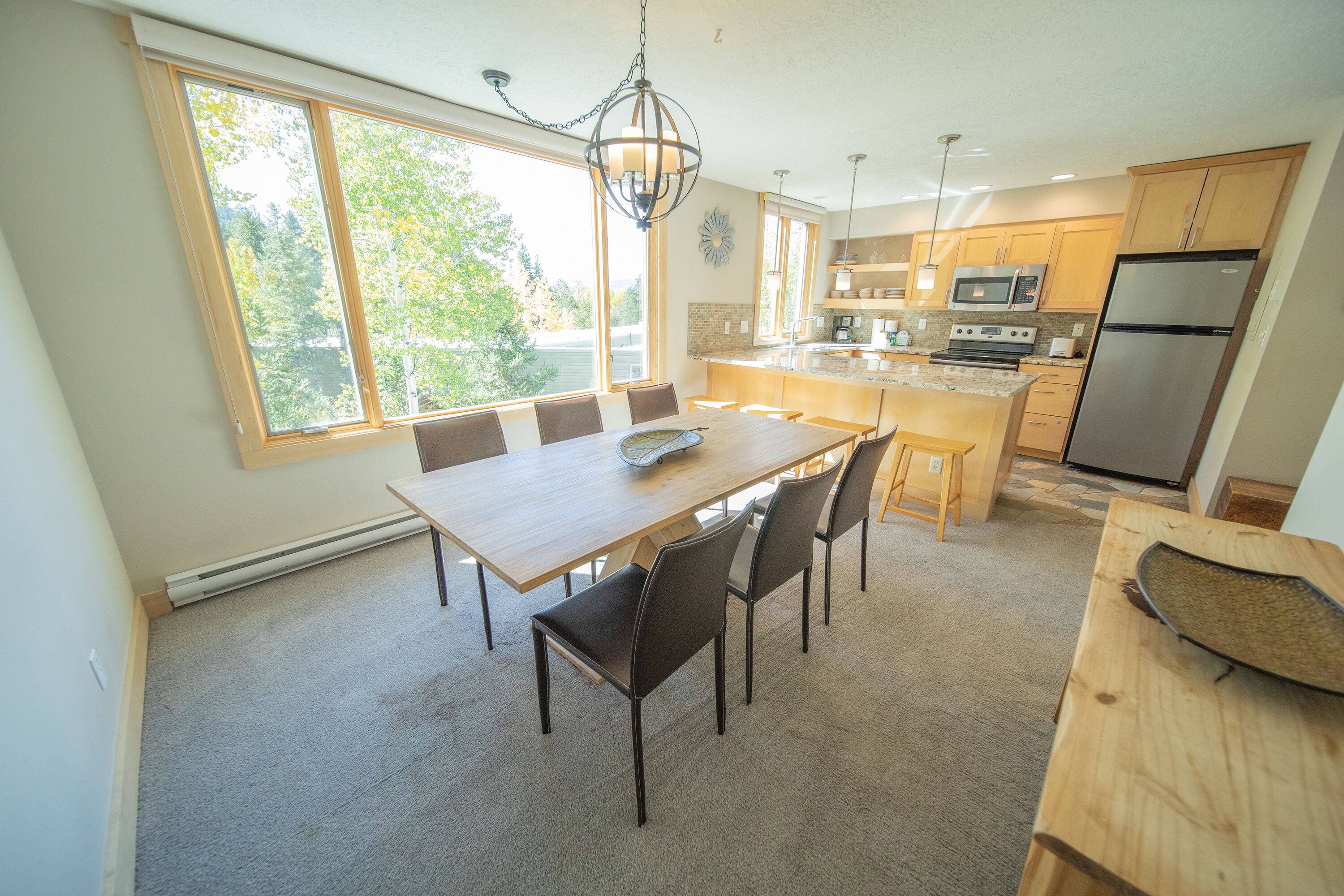 A modern kitchen with a dining area, large windows, and a view of trees.