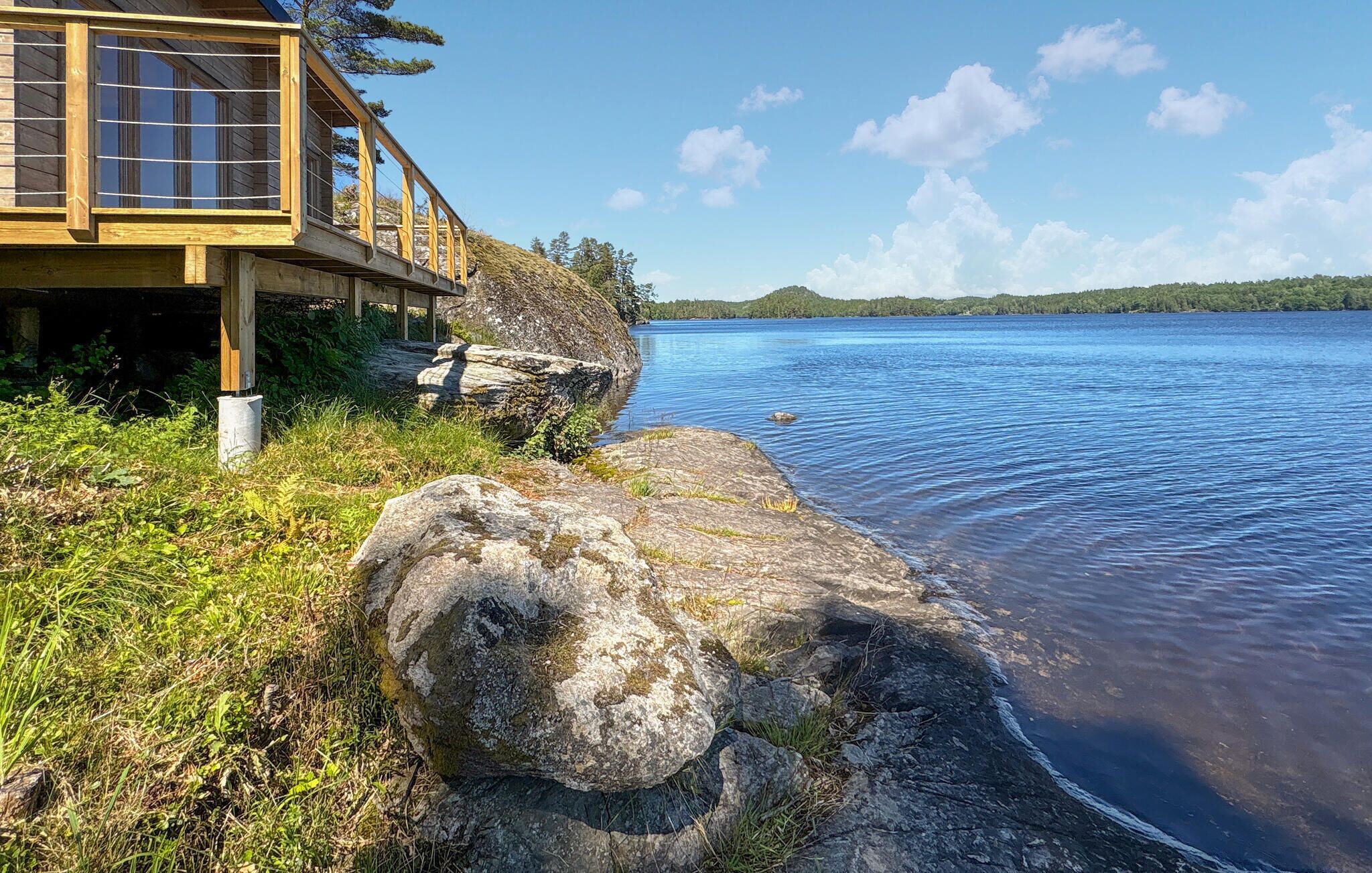 A wooden cabin with a deck overlooking a lake, surrounded by rocks and greenery.