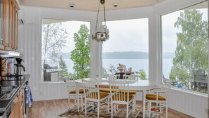 A dining area with a round table and white chairs, large windows with a view of a lake, and a hanging light fixture. - Lovely home in Färgelanda with sauna (Färgelanda)