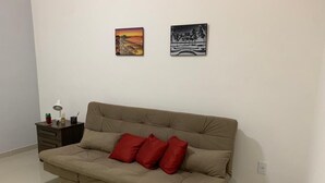 En beige sofa med fire røde puder, et lille sengebord med en lampe, og to billeder hængende på væggen.