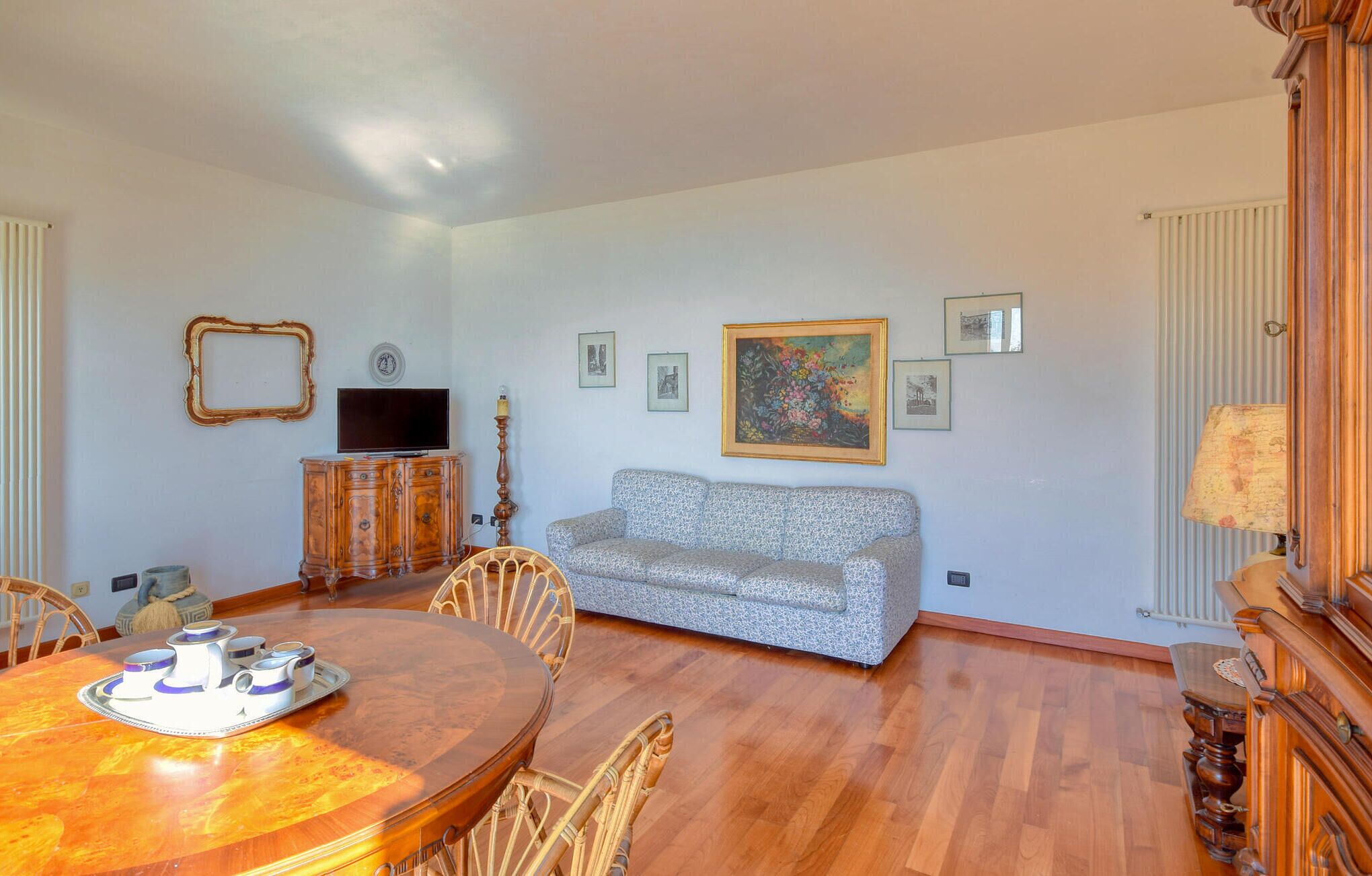 A living room with a wooden dining table, wicker chairs, a grey sofa, a television, and framed pictures on the wall.