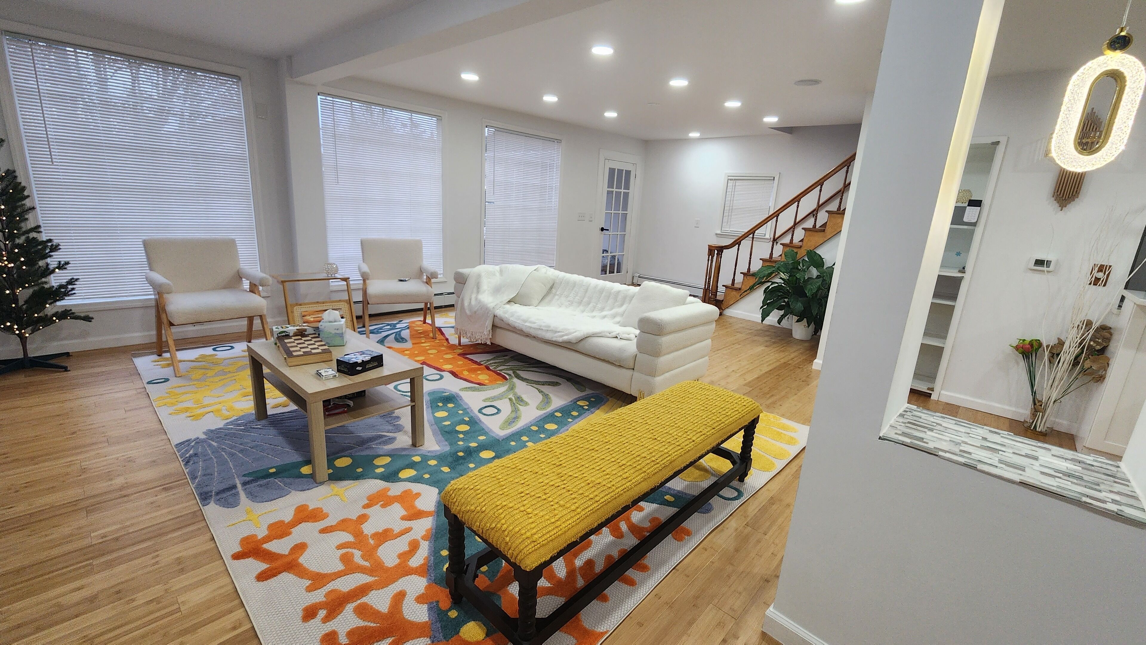 A spacious living room with a white sofa, two armchairs, a coffee table, and a bench. There is a staircase leading to another level, a small Christmas tree, and a potted plant.
