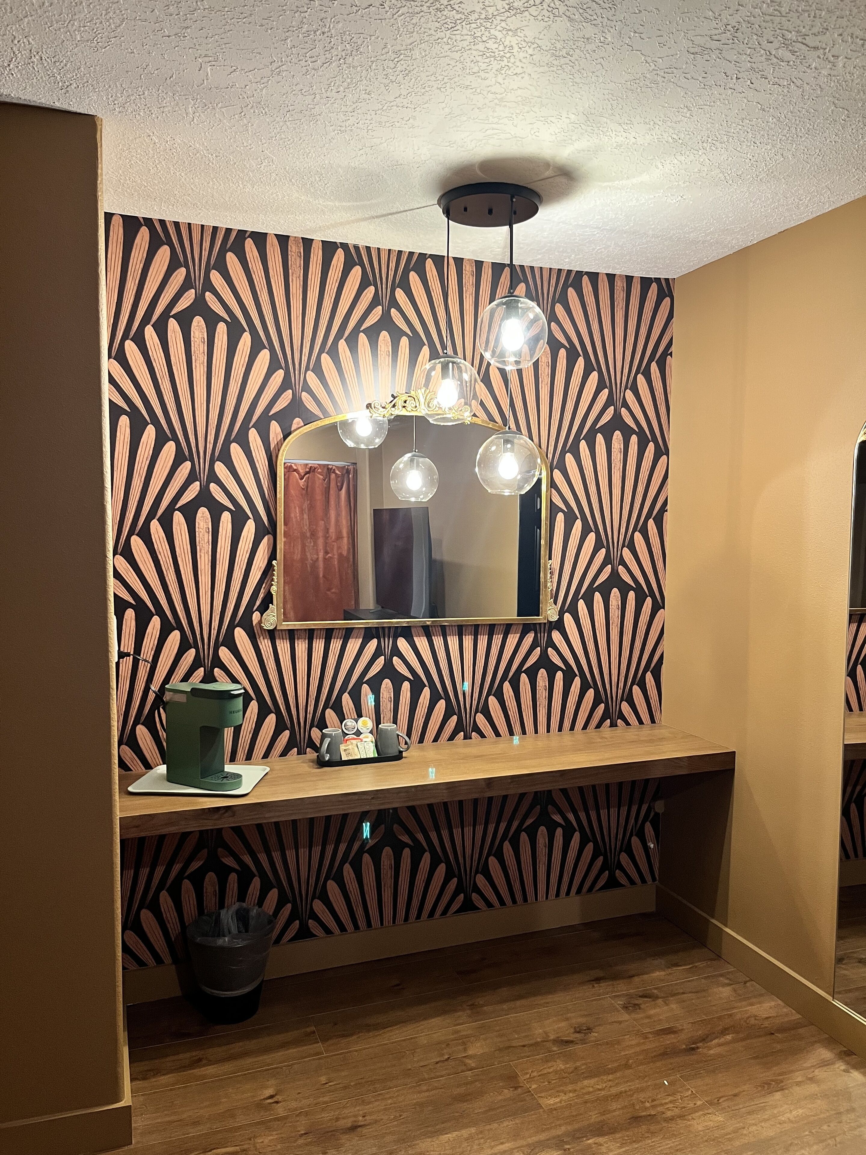 Une salle de bain avec un mur à motifs, un miroir, un meuble-lavabo avec une machine à café et un luminaire au plafond.