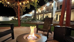 An outdoor seating area with a circular fire pit, wooden chairs, and a covered patio with string lights. - El Verde Inn (Albuquerque)