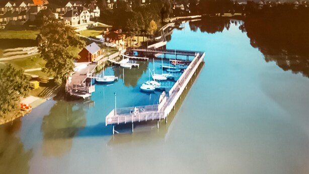 Vue aérienne d’une marina où sont amarrés plusieurs bateaux, avec une jetée en bois et des immeubles résidentiels environnants.