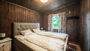 Een slaapkamer met een wand van houten panelen, een gestoffeerd hoofdbord, een bed met een beddegoed met een patroon, een nachtkastje met een lamp, en een raam met gordijnen.