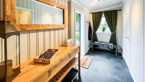 Een kamer met een houten bankje, een spiegel, een raam met gordijnen, een deurmat en een klein tafeltje met een doos en een bak.