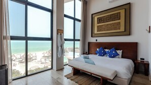 A bedroom with a large window overlooking a beach, a bed with blue pillows, a wooden headboard, and a framed artwork on the wall.