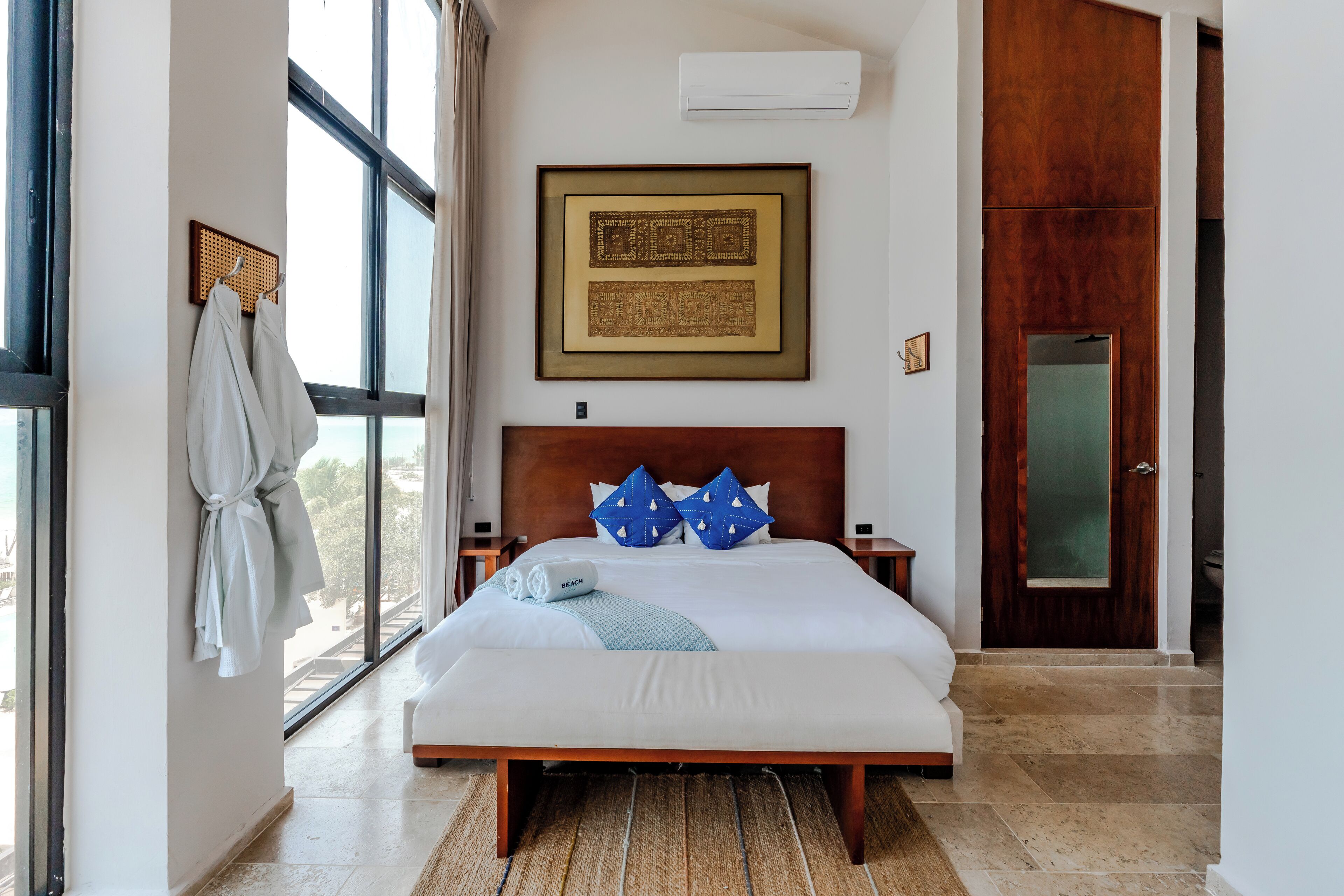 A modern hotel room with a large bed, a wooden headboard, a framed artwork on the wall, and a view through the window.
