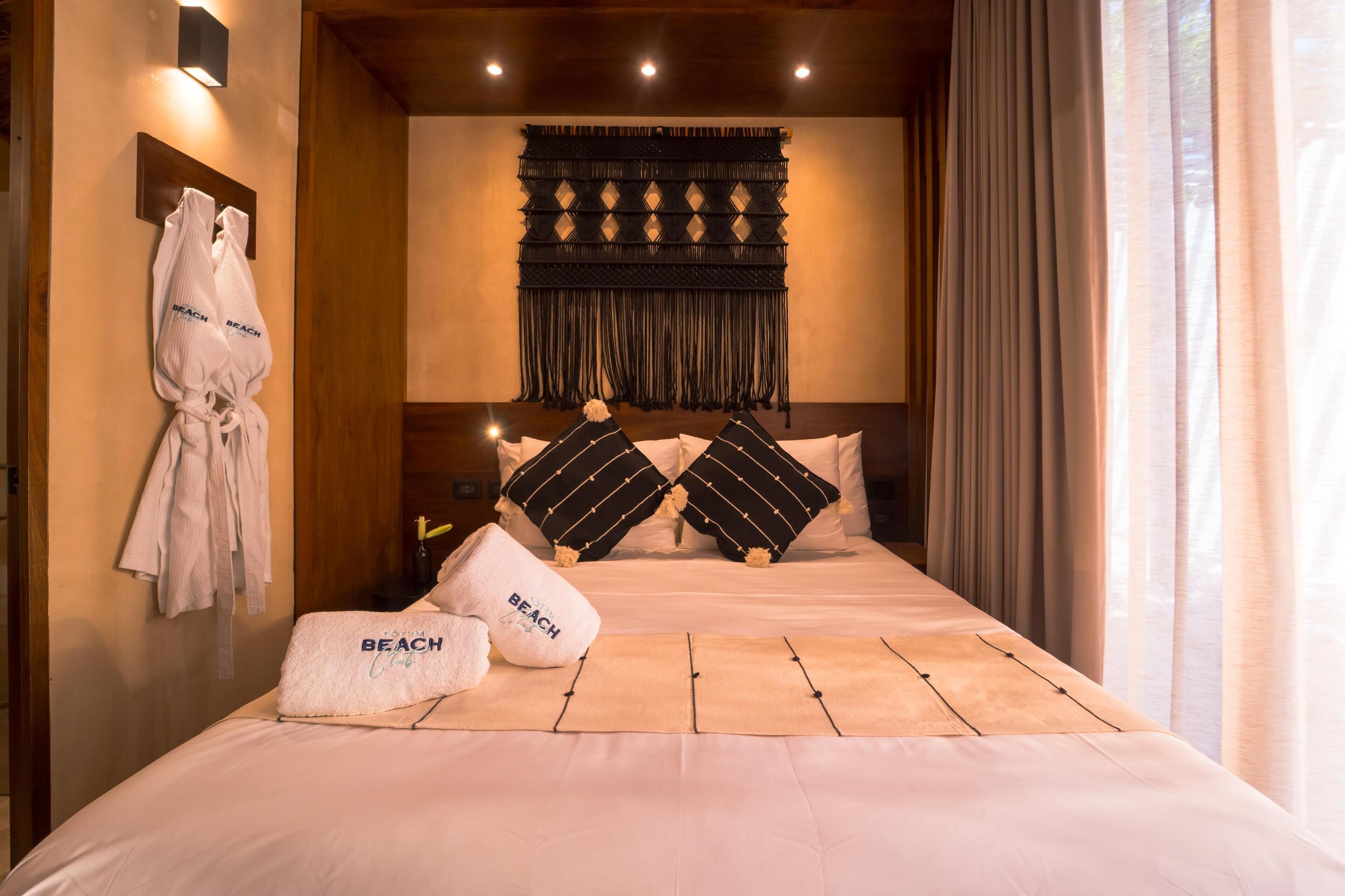 A hotel room with a large bed, two pillows, a wall-mounted towel rack with towels, and a decorative wall hanging.