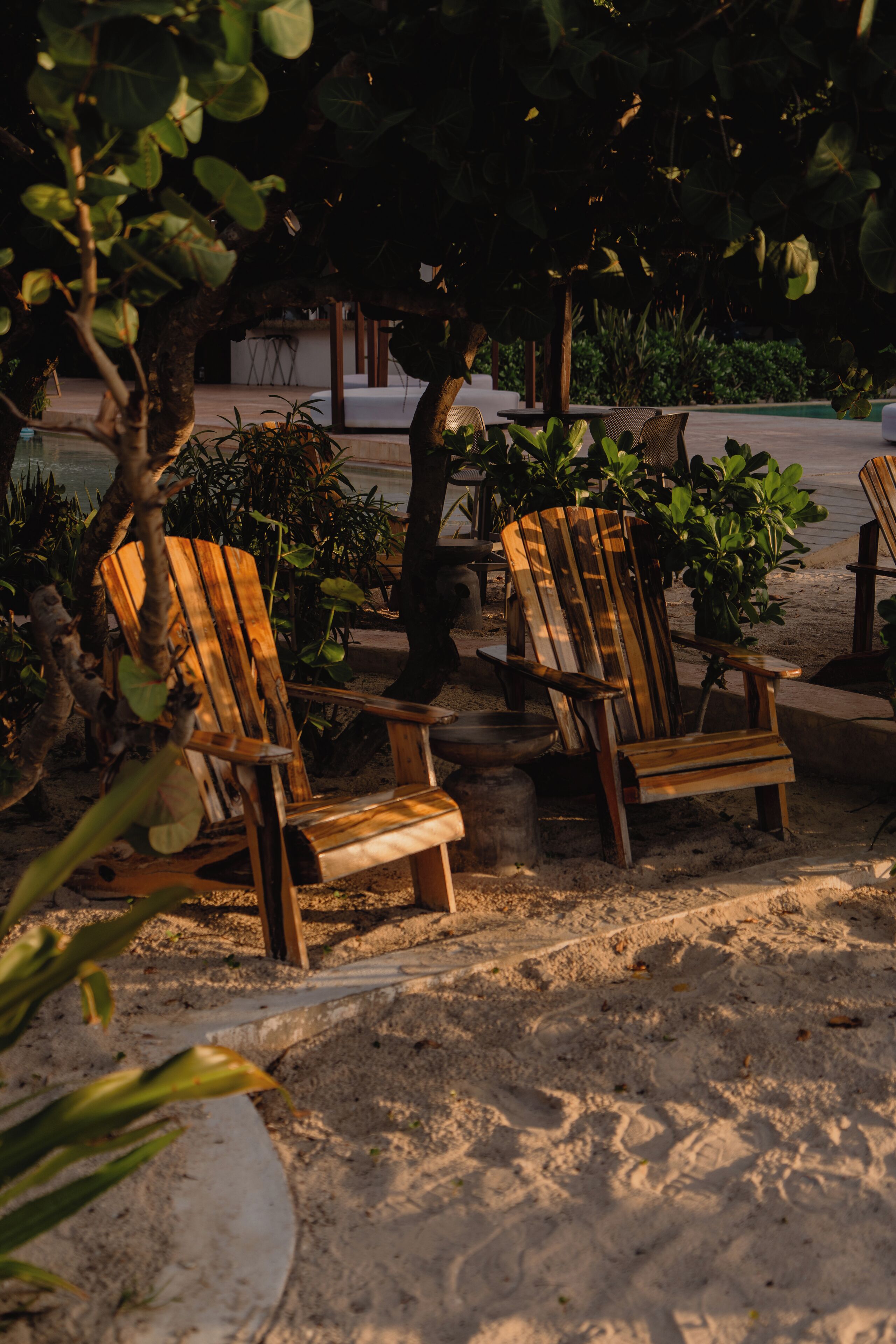 Outdoor wooden chairs with a sandy ground, surrounded by greenery and a building in the background.