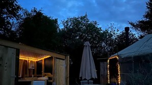 Ulkokäyttöön tarkoitettu oleskelualue, jossa on puinen pöytä ja tuolit, pieni puinen rakennelma, jossa on valonlähde, sekä katettu aurinkovarjo.