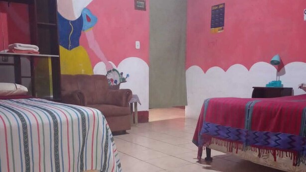Una habitación con dos camas, una silla marrón, una mesa con una lámpara y un mural en la pared.