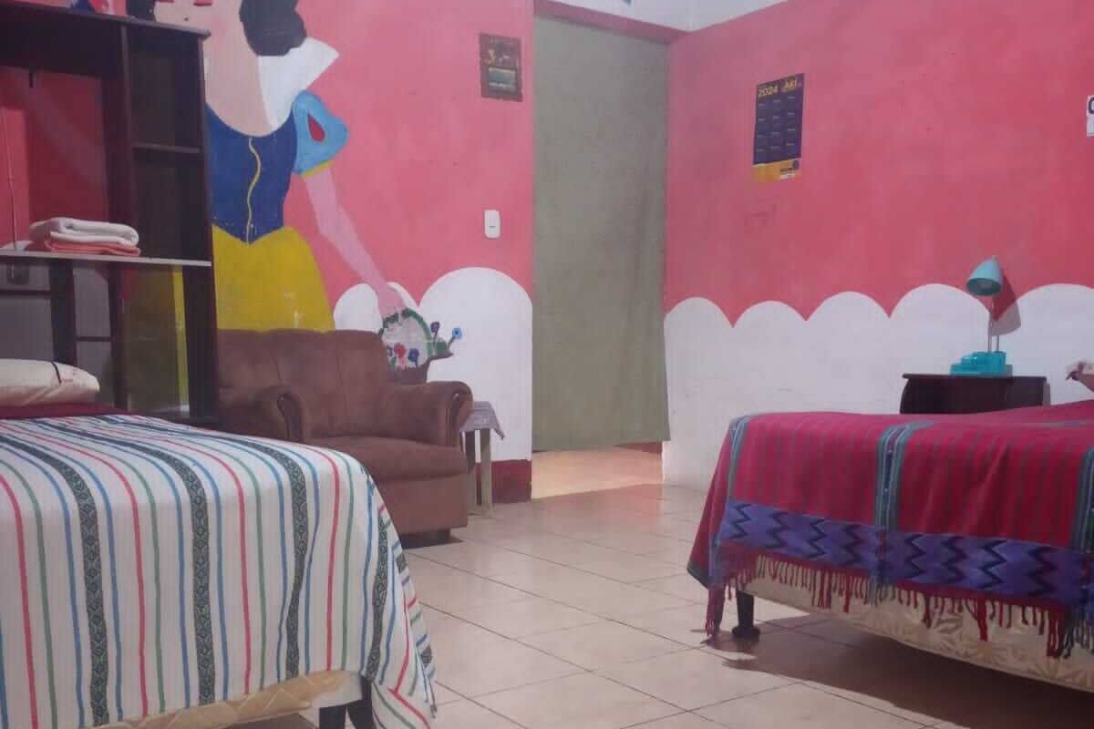 A room with two beds, a brown chair, a table with a lamp, and a wall mural.