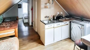 Cocina compacta con armarios blancos, un lavabo y una tetera. Hay una pequeña zona de comedor con una mesa y una silla.