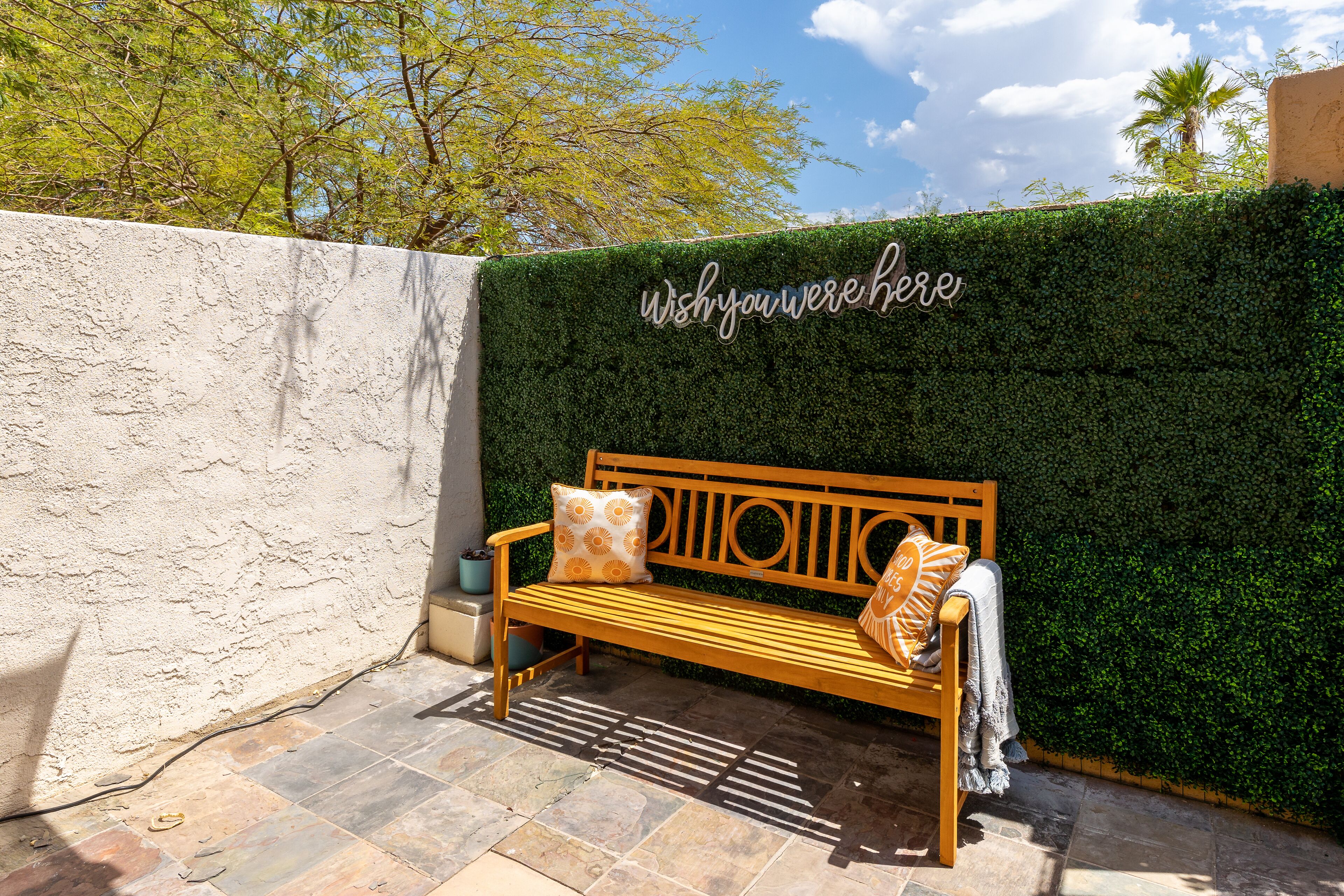 A wooden bench with patterned cushions against a green hedge wall with a sign that reads "Wish you were here."