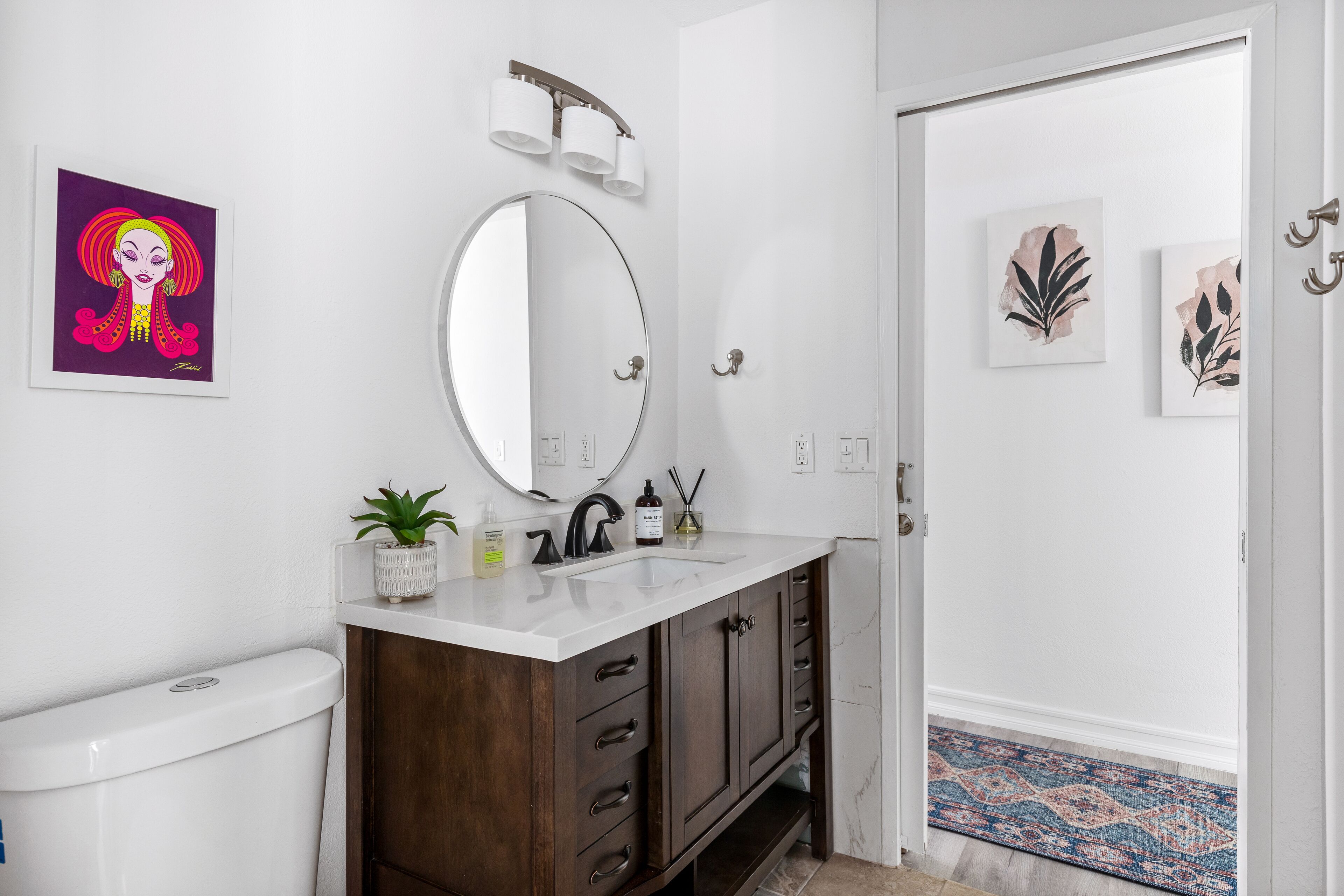 A bathroom with a white toilet, a wooden vanity with a mirror, and a sink. There are wall-mounted lights, a potted plant, and a decorative piece on the wall.