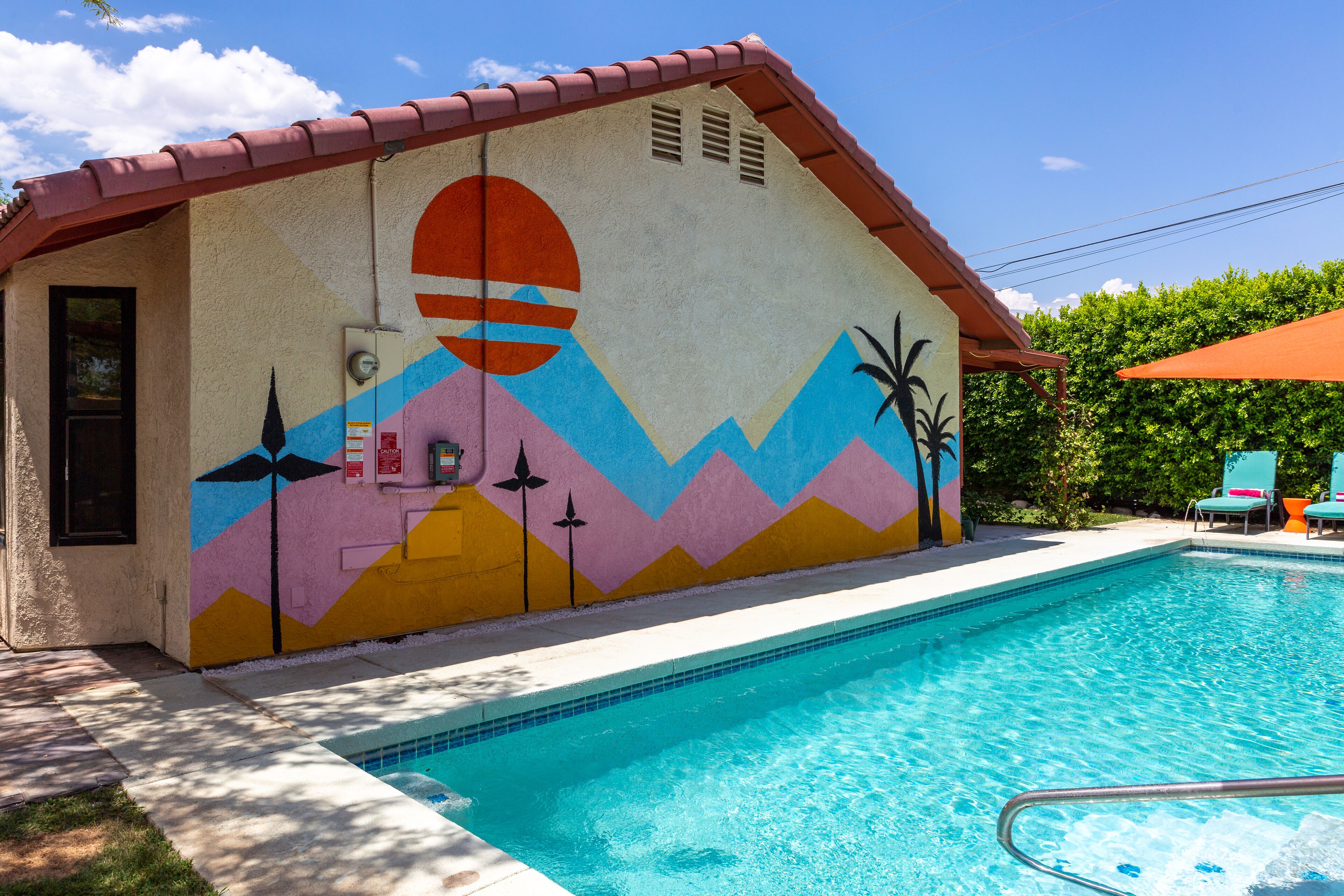 A swimming pool with a colorful mural on the adjacent wall, a clear blue sky, and a red umbrella.