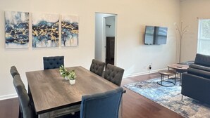 A spacious living room with a dining area, featuring a wooden dining table, blue upholstered chairs, a ceiling fan, and a flat-screen TV mounted on the wall.