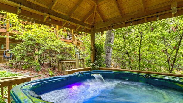 A hot tub under a wooden pergola with a view of a wooden deck and lush greenery.