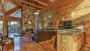 L’intérieur d’une cabane en rondins, avec une cheminée en pierre, des poutres en bois au plafond, un canapé en cuir et un coin cuisine.