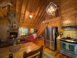 A cozy cabin interior with a fireplace, wooden dining table, kitchen with stainless steel appliances, and a staircase leading to a loft area.