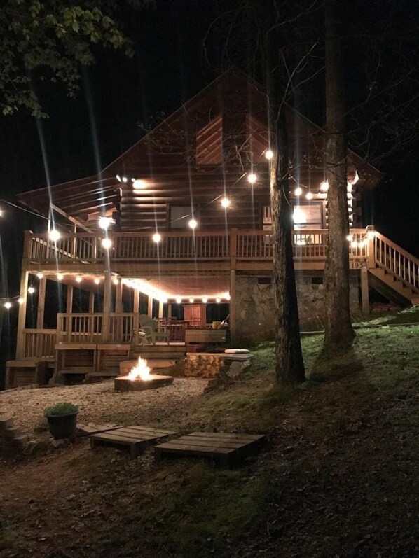 A wooden cabin with a deck, string lights, and a fire pit.