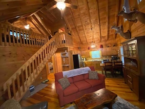 A wooden cabin interior with a red sofa, wooden staircase, kitchen area, and a ceiling fan.