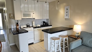 Ett kompakt kök med vita skåp, en mikrovågsugn, en spis och en kaffebryggare. Det finns två barstolar och ett bord med en lampa.