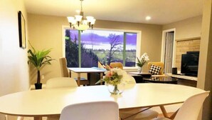 Un coin repas avec une table ronde, des chaises blanches, un lustre et une fenêtre donnant sur des arbres et un ciel violet.
