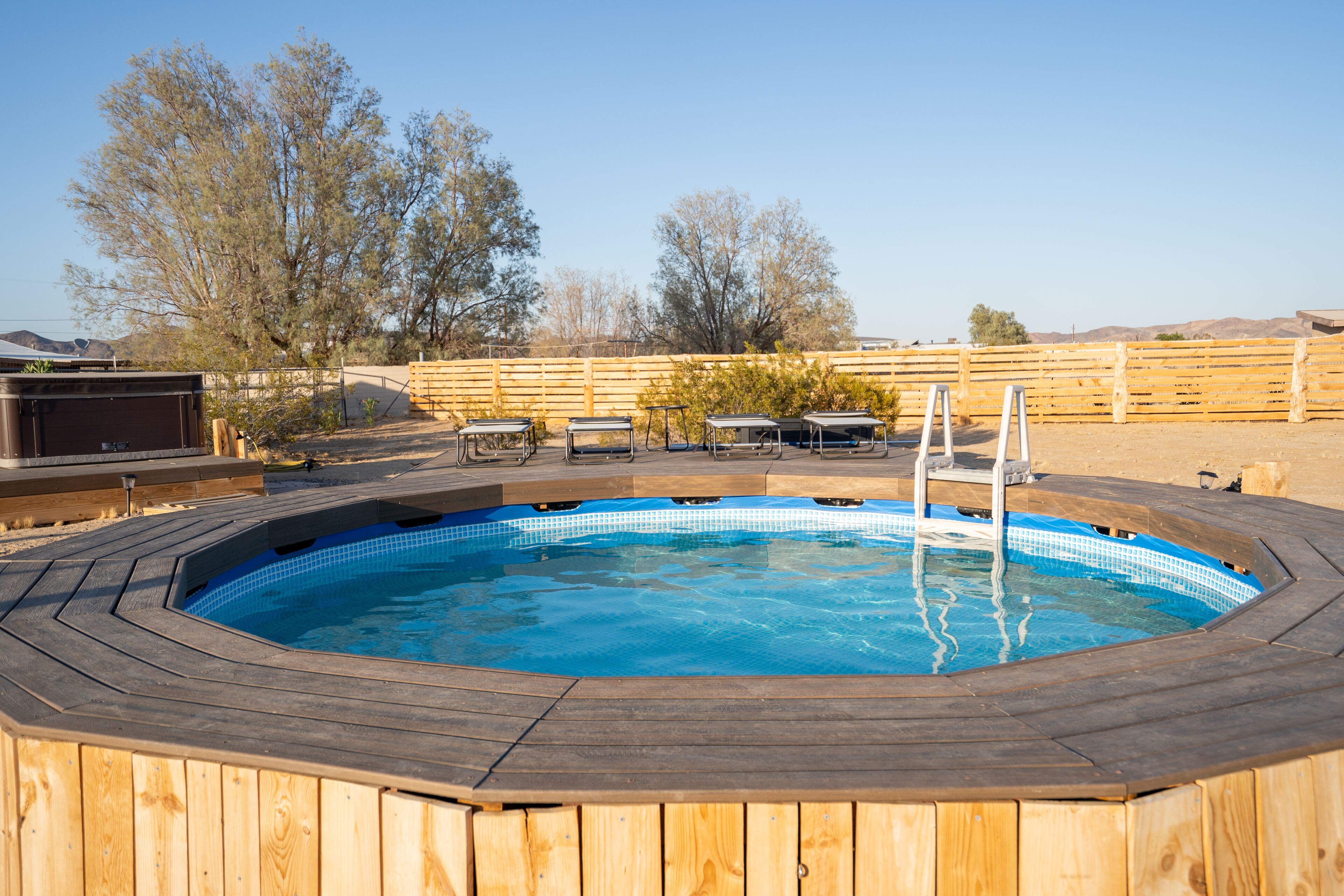 A wooden deck with a hot tub, surrounded by a wooden fence, trees, and a clear sky.