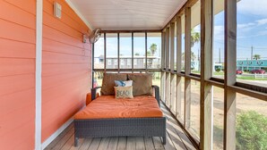 En inglasad veranda med en flätad säng, en prydnadskudde med texten ”BEACH” och utsikt över ett bostadsområde med palmer och en blå byggnad.
