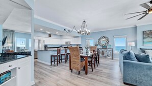 A modern kitchen with white cabinets, a dining area with wicker chairs, and a view of the ocean. - Admirals Quarters 1101 | Gulf-Front Views | Beach Getaways (Orange Beach)