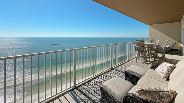 A balcony with a view of the ocean, a sofa with patterned cushions, and a table with chairs. - Crystal Shores West 1402 | Beachfront Penthouse | Pools, Fitness, Sauna | Beach Getaways (Gulf Shores)