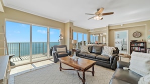 A spacious living room with a large window overlooking the ocean, a ceiling fan, and a cozy seating area with a coffee table. - Crystal Shores West 1402 | Beachfront Penthouse | Pools, Fitness, Sauna | Beach Getaways (Gulf Shores)