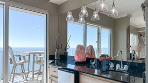 A modern kitchen with a large window, a black countertop, and a view of the ocean. - Crystal Shores West 1402 | Beachfront Penthouse | Pools, Fitness, Sauna | Beach Getaways (Gulf Shores)