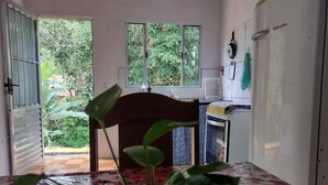 Ein Zimmer mit einem Esstisch, der mit einer blumigen Tischdecke bedeckt ist, einer Zimmerpflanze in der Mitte und Blick ins Grüne durch die offene Tür.
