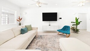 A modern living room with a flat-screen TV, a beige sofa, a blue rocking chair, and a white cabinet. - Moonflower Villa: Luxe Oasis in Sunset Crest, Holetown (Sunset Crest)
