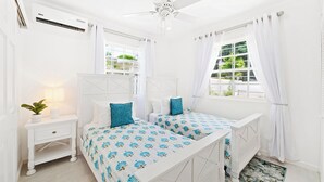 Une chambre avec deux lits, un ventilateur de plafond, un appareil de climatisation, une table de chevet avec une lampe et une fenêtre avec des rideaux.