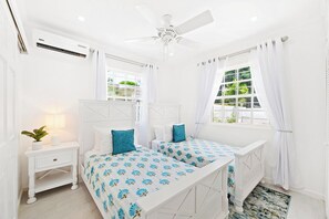 A bedroom with two beds, a ceiling fan, an air conditioning unit, a bedside table with a lamp, and a window with curtains. - Moonflower Villa: Luxe Oasis in Sunset Crest, Holetown (Sunset Crest)