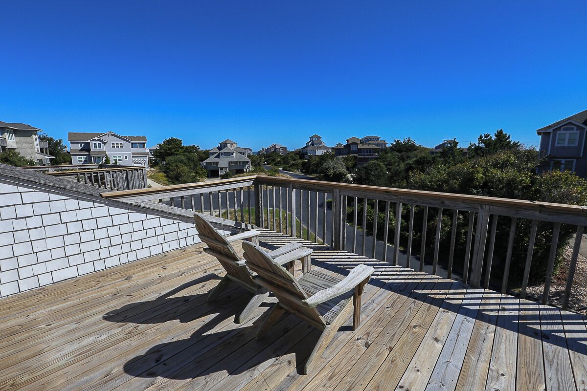 En træterrasse med Adirondack-stole og et gelænder i træ, med udsigt over et boligområde med huse og træer.