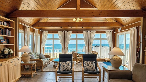 Une pièce spacieuse avec un plafond haut, des poutres en bois, de grandes fenêtres avec des rideaux, un canapé, des fauteuils, une table et une bibliothèque.