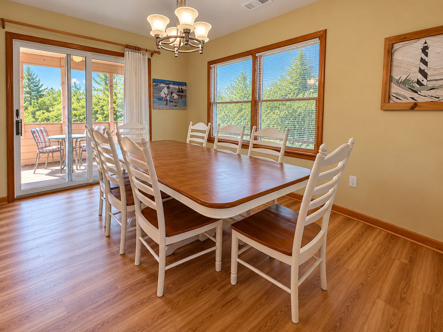 A dining room with a wooden table and white chairs, large windows with blinds, and a chandelier.