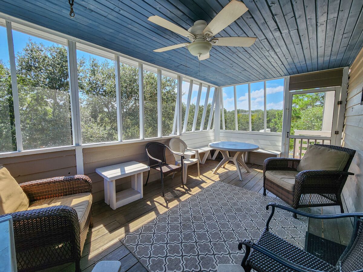 A covered patio with wicker furniture, a ceiling fan, and large windows offering views of trees.