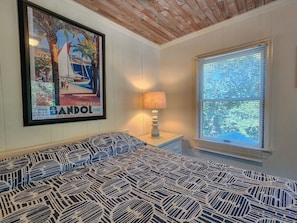 A bedroom with a patterned bedspread, a framed picture on the wall, a bedside lamp, and a window with blinds.